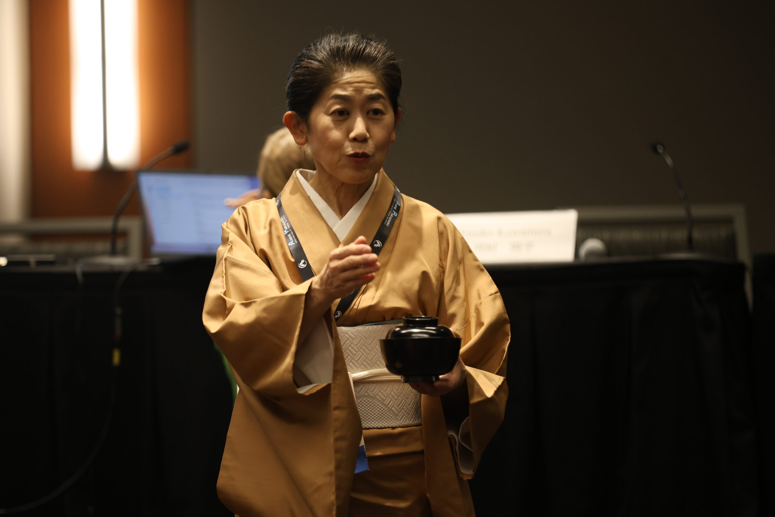 A Japanese woman wearing a kimono speaks in front of a room. In her left hand, she holds a Japanese style bowl.